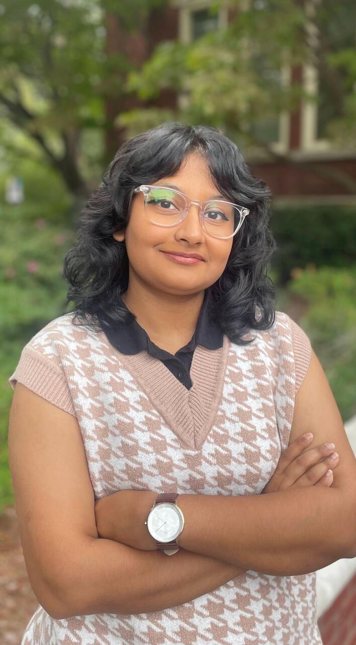 Shirley's Headshot A picture of Shirley wearing a sweater vest and a brown watch, crossing his arms with trees in the background.