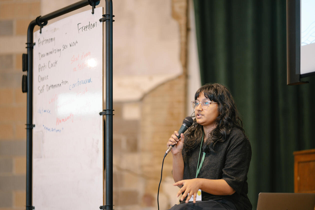 Shirley leading a workshop on Crip Solidarity Shirley speaking on a microphone next to a whiteboard with hand-written words reading: Autonomy, Decriminalizing sex work, Anti-caste, Choice, Freedom, Self-Determination, Humanity, continuum